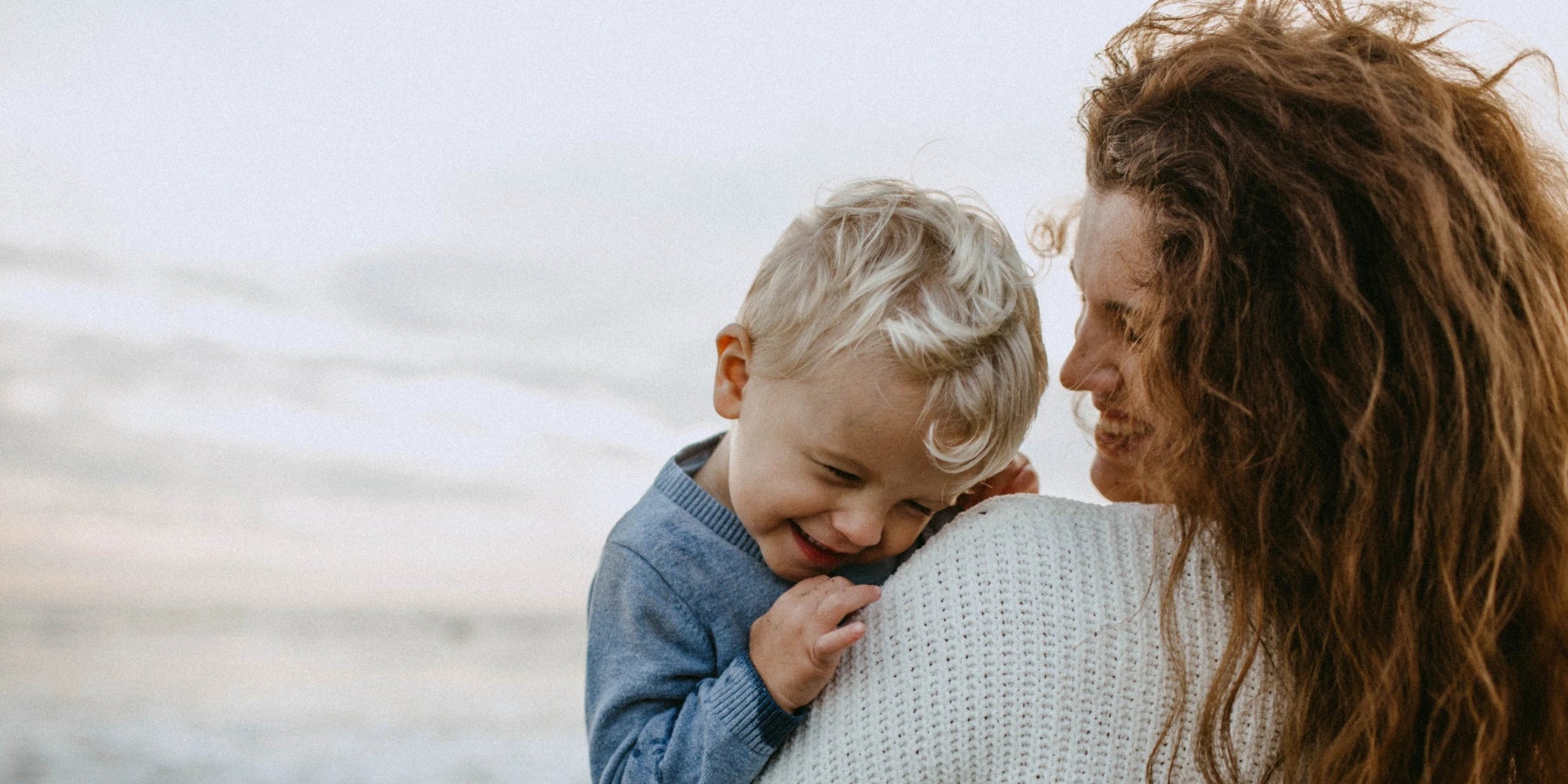 Mutter hält lachenden kleinen Sohn am Strand im Arm – inniger Familienmoment, Symbol für Mutterliebe, Geborgenheit und Geschenkideen für Mamas und Kinder von Amorlune